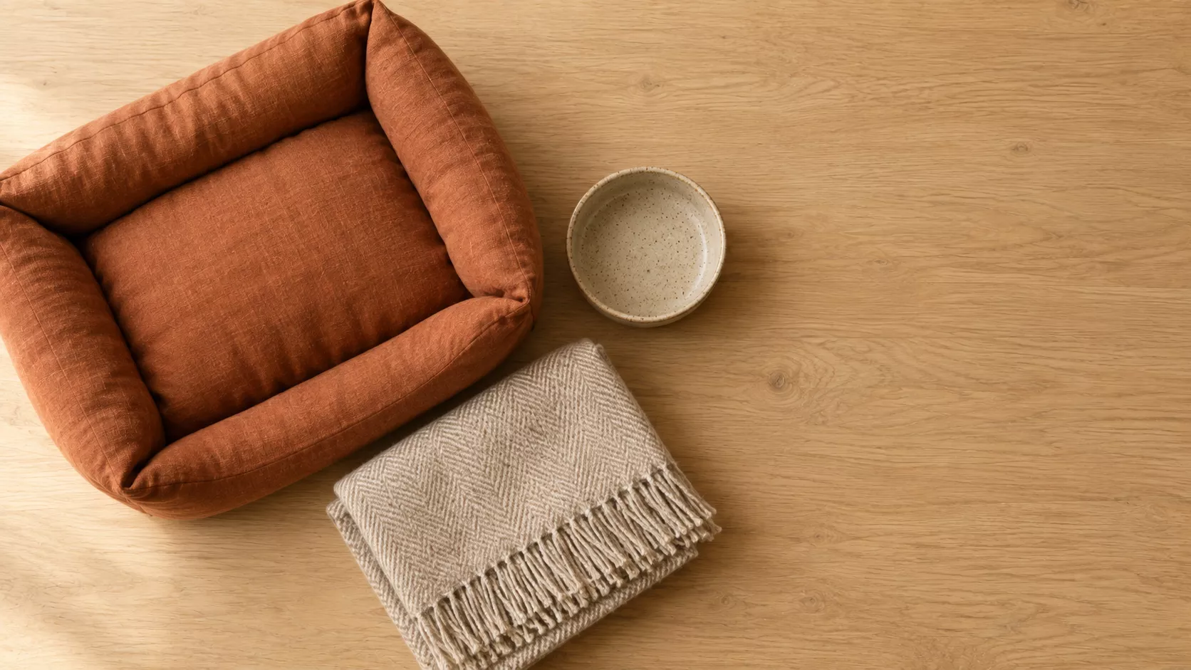 A terracotta linen dog bed, a speckled stoneware bowl and a fringed wool throw on an oak floor